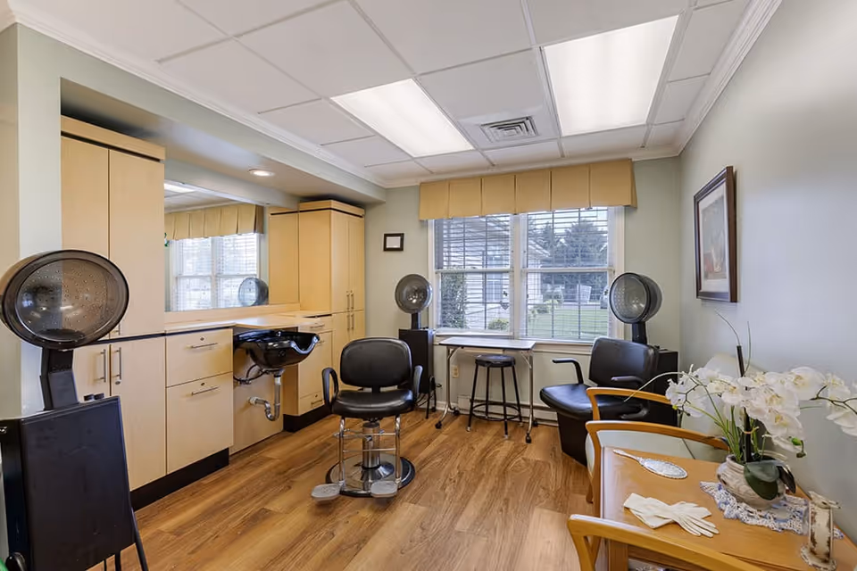 Interior of a hair salon area within a senior living facility, featuring two black salon chairs with hair dryers, a black sink for washing hair, wooden cabinets, a large mirror, a window with a beige valance, a small table with a stool, and a wooden bench with white gloves and a vase of white flowers on it.