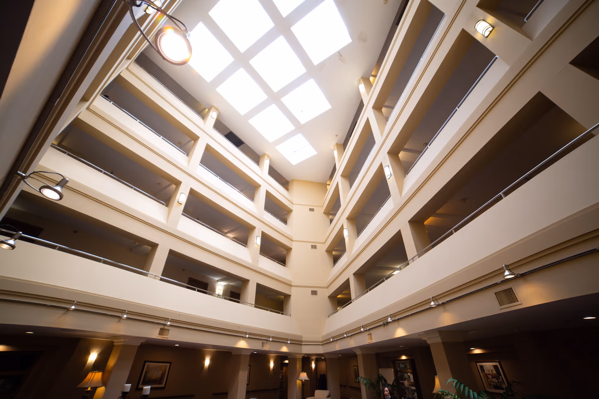 Interior view of a multi-story building atrium with beige walls and railings on each floor. The ceiling features large skylights allowing natural light to illuminate the space. Wall-mounted lights and ceiling spotlights provide additional lighting. The lower level has some furniture and framed artwork on the walls.