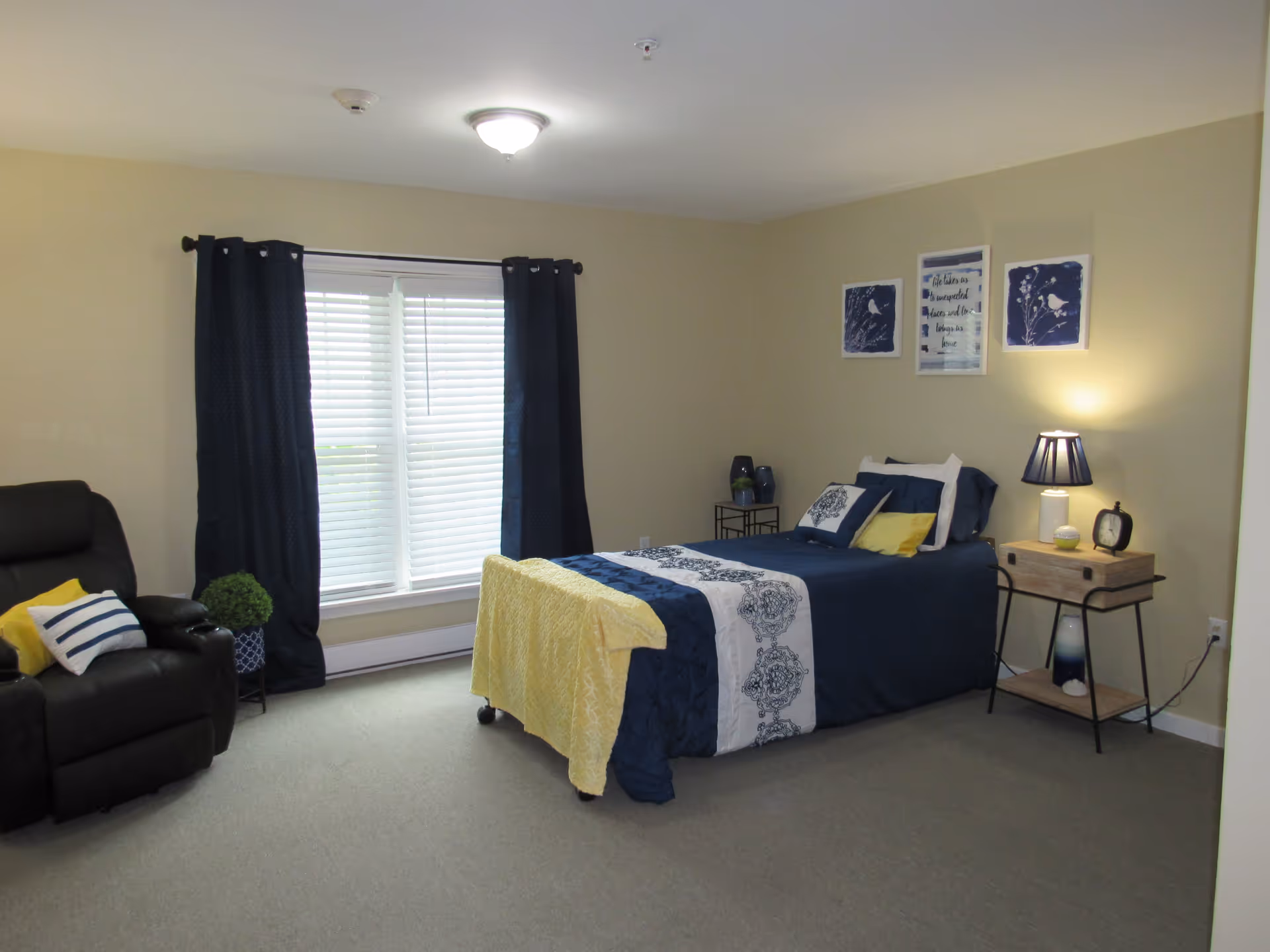 A bedroom with a single bed dressed in navy blue and white bedding with a yellow throw blanket. The room has beige walls, a window with white blinds and navy blue curtains, a black recliner chair with yellow and striped pillows, a small plant in a blue pot, and a wooden nightstand with a lamp, clock, and decorative items. Three framed pictures hang on the wall above the bed.