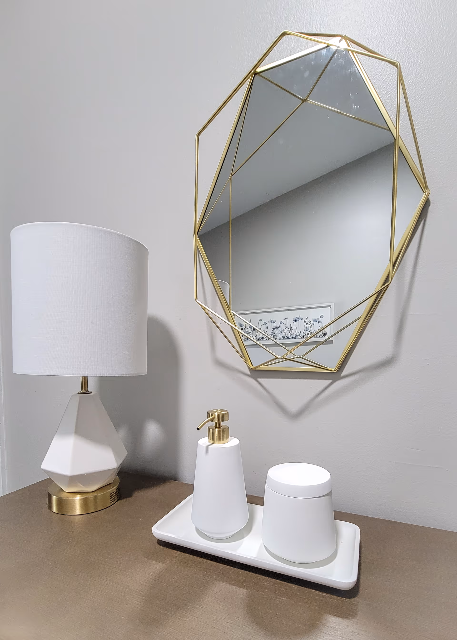 A modern vanity setup with a geometric gold-framed mirror mounted on a light gray wall. Below the mirror is a brown countertop holding a white geometric table lamp with a gold base, and a white tray with a white soap dispenser and a white container with a lid.