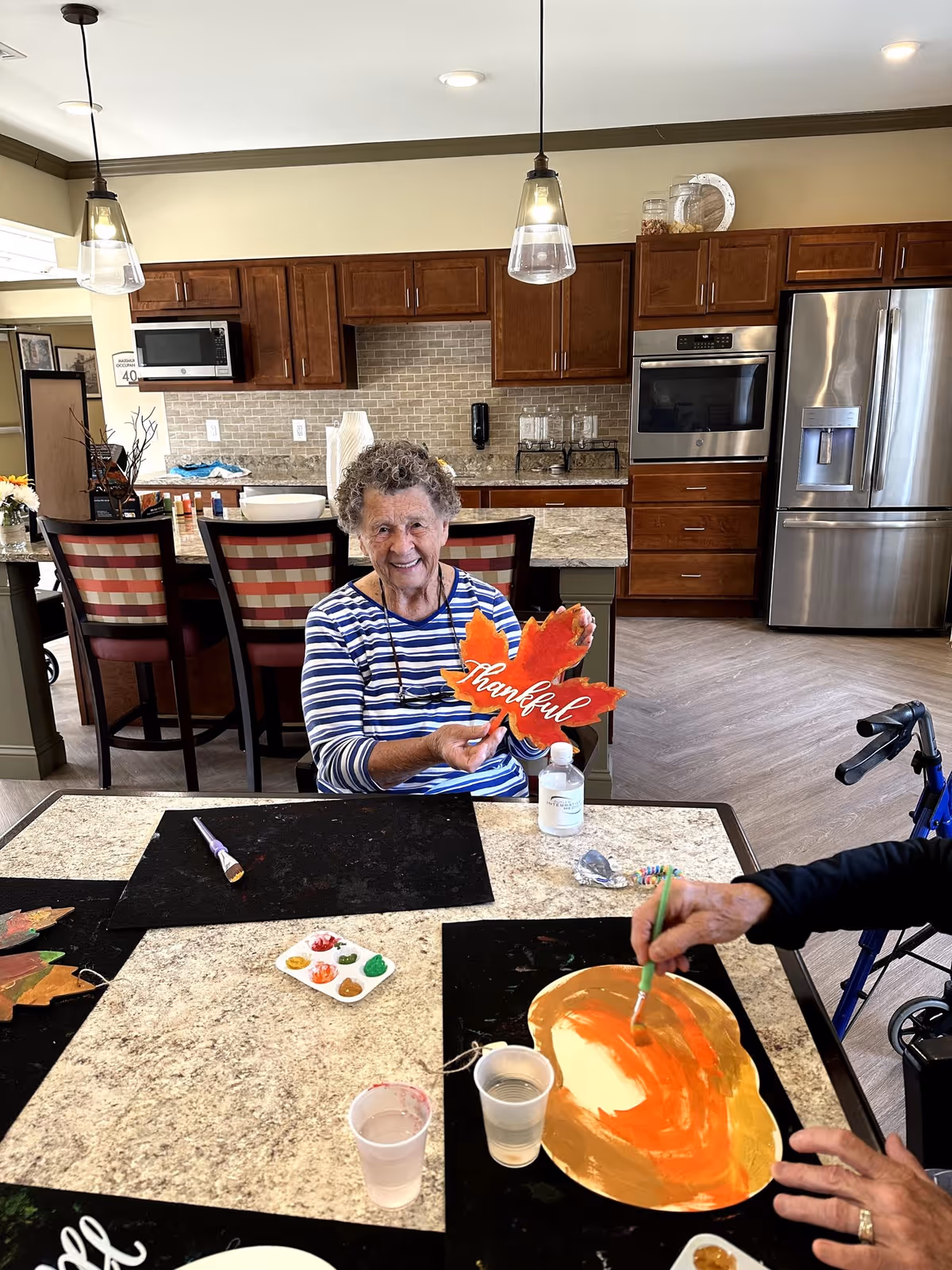 An elderly woman sitting at a table in a kitchen area, holding a decorative orange leaf with the word 'thankful' written on it. Another person is painting a pumpkin-shaped craft with orange paint. The kitchen has wooden cabinets, a stainless steel refrigerator, and pendant lights hanging from the ceiling.