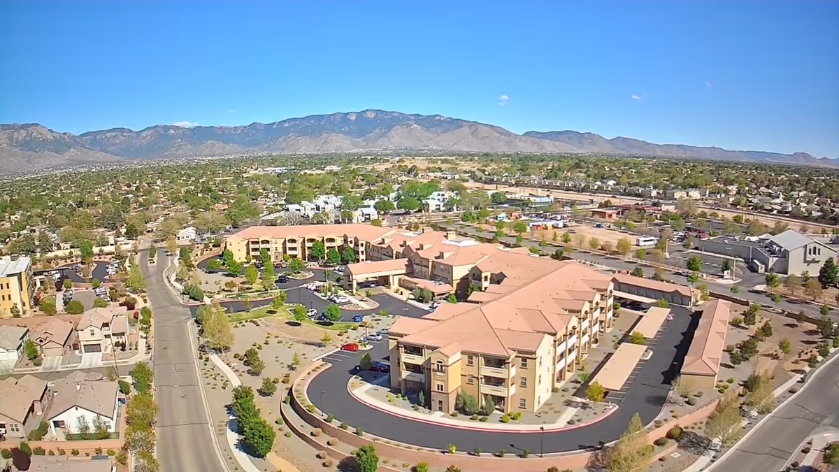 Aerial view of the Coronado Villa Retirement Resort showing tan multi-story buildings, parking areas, surrounding neighborhood and distant mountains under a clear sky.