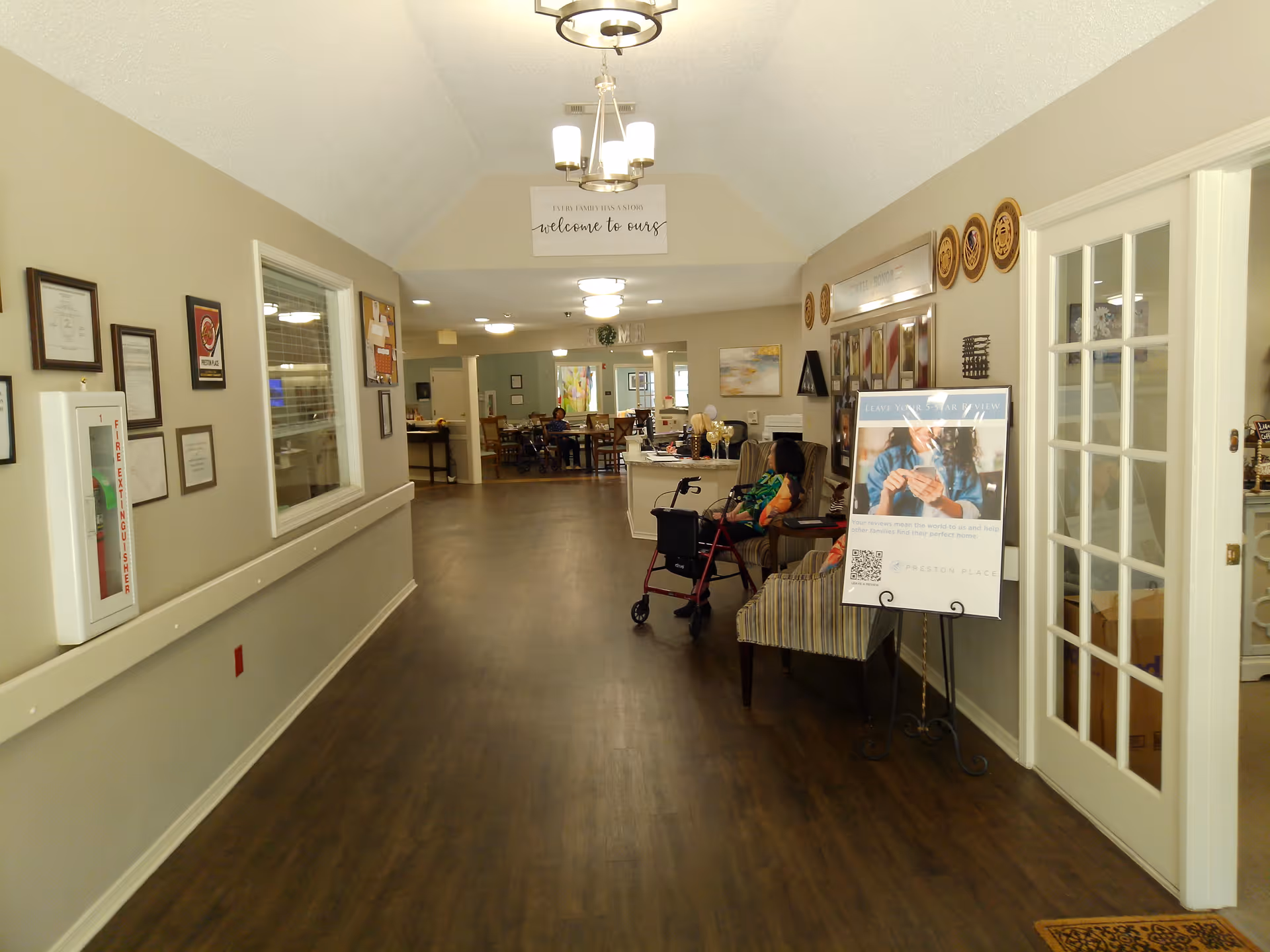 Interior hallway of a senior living facility with wood flooring, beige walls, and ceiling lights. On the right side, there is a woman sitting in a chair next to a walker, and a signboard with information. The hallway leads to a dining area with tables and chairs where a few people are seated. Various framed pictures and certificates are displayed on the walls.
