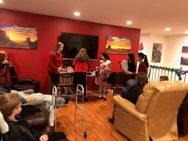 A group of visitors and residents gathered in a senior living room with a TV, bookshelves, recliners, and holiday decorations.