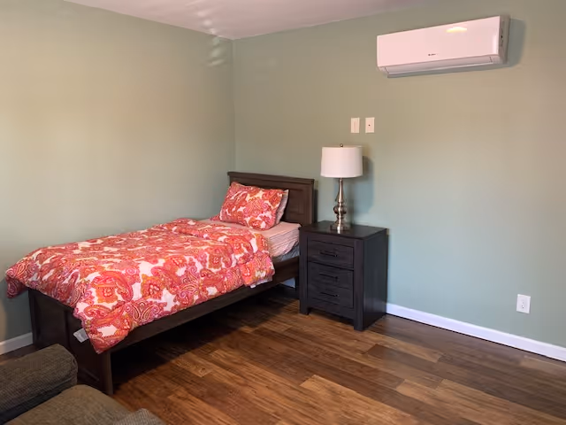 A simple bedroom with a single bed covered in a red and pink patterned comforter and pillow. Next to the bed is a dark wooden nightstand with a white lamp on it. The walls are painted light green, and there is a white air conditioning unit mounted on the wall above the nightstand. The floor is wooden, and part of a dark-colored chair is visible in the foreground.