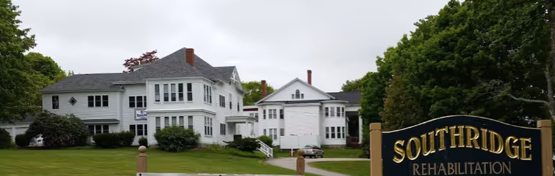 Exterior view of Southridge Rehabilitation facility showing two large white multi-story buildings surrounded by green trees and bushes under a cloudy sky. A black and gold sign with the text 'SOUTHRIDGE REHABILITATION' is visible in the foreground on the right side.
