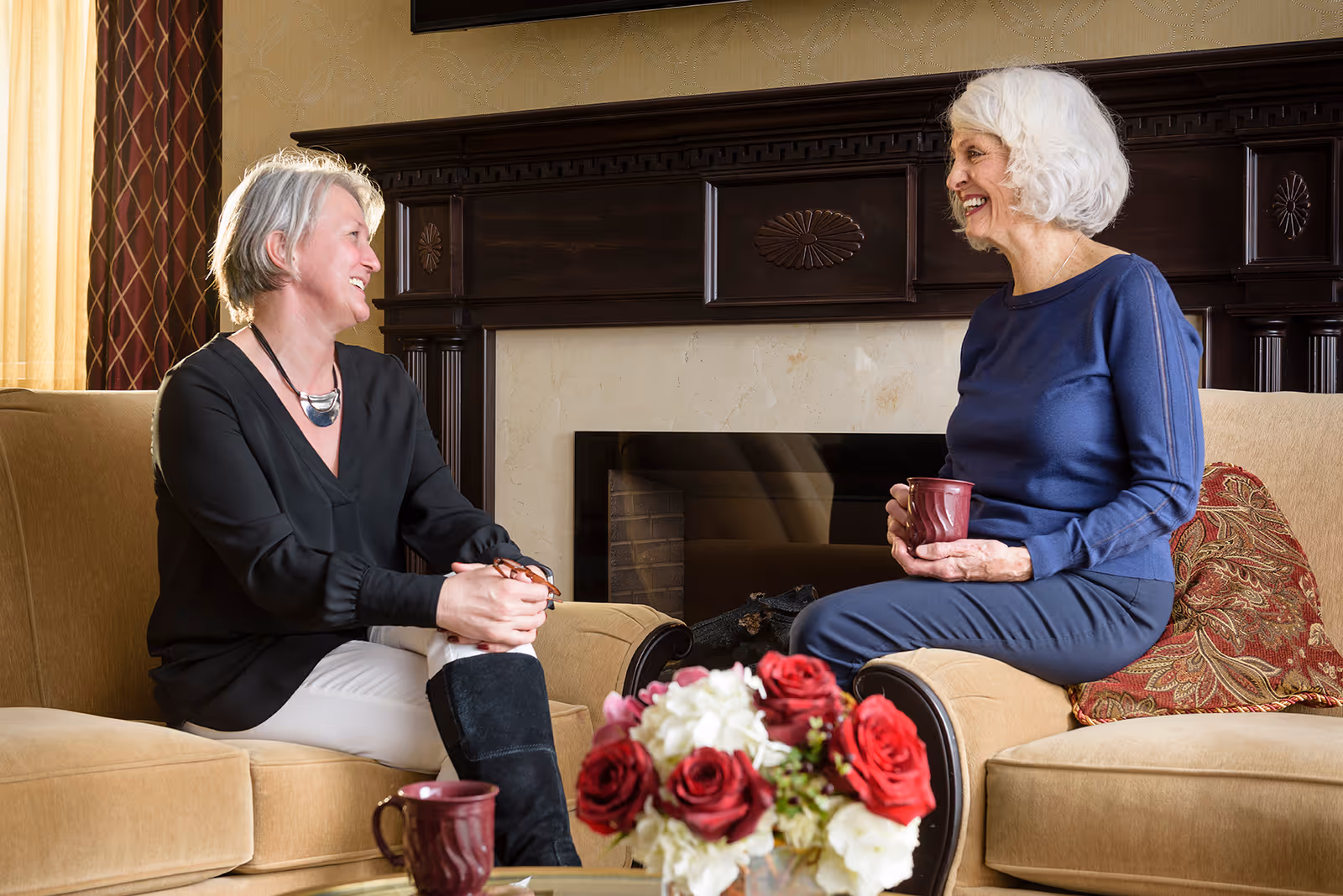 Two elderly women sitting on beige sofas in a cozy living room, smiling and chatting. One woman has short gray hair and is wearing a black blouse and white pants, while the other has white hair and is wearing a blue top and pants. They each hold a maroon mug. A bouquet of red and white flowers is on the glass coffee table in front of them, and a dark wooden fireplace is in the background.