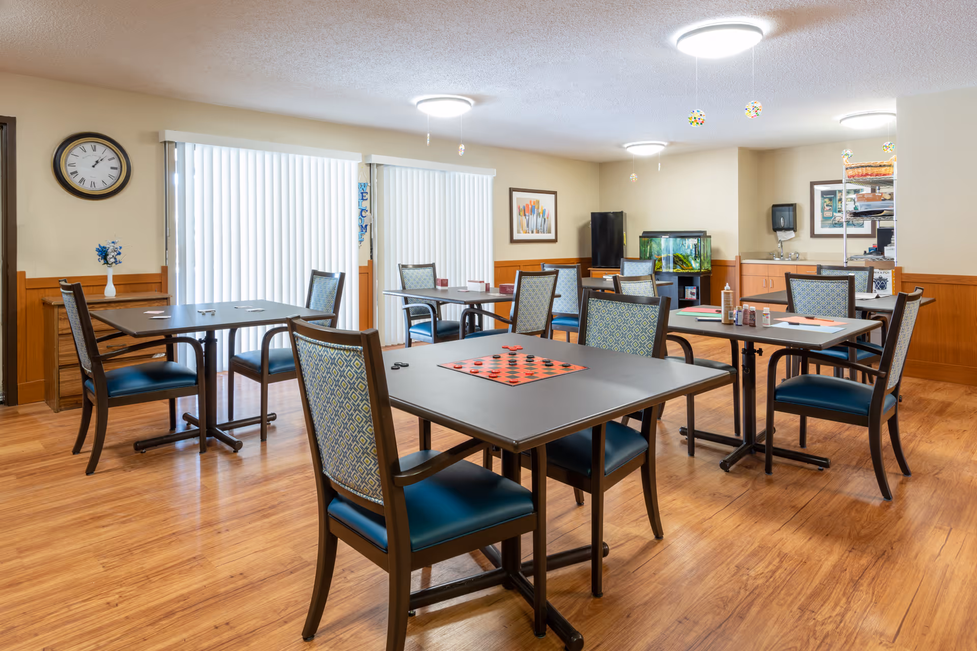 A bright and clean common room with several tables and chairs arranged for group activities. One table has a checkers board set up, and other tables have paper and markers. The room has wood flooring, beige walls with wood paneling, a clock on the wall, a fish tank, and a small sink area with a paper towel dispenser. Vertical blinds cover the windows, and ceiling lights illuminate the space.