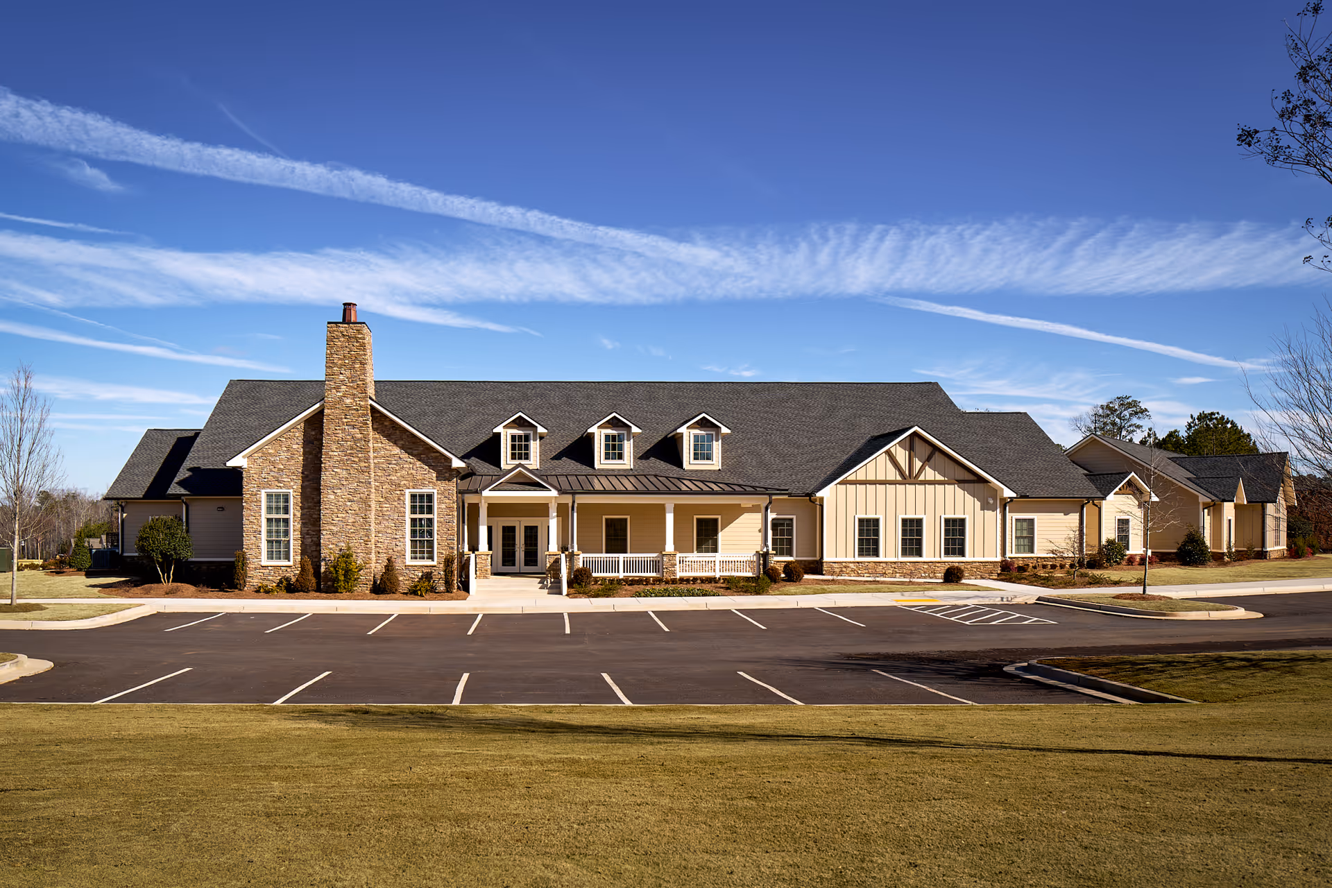 Front exterior view of a single-story senior living facility building with a stone chimney, multiple windows, a covered porch entrance, and a large empty parking lot in front under a clear blue sky.