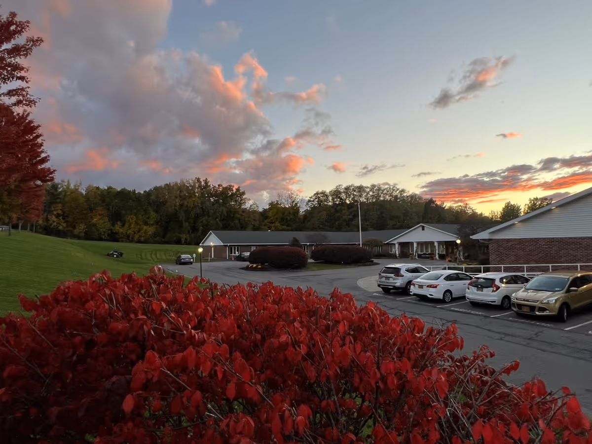 View of a senior living facility parking lot at sunset with several parked cars, red bushes in the foreground, green grass and trees in the background, and a partly cloudy sky with pink and orange hues.