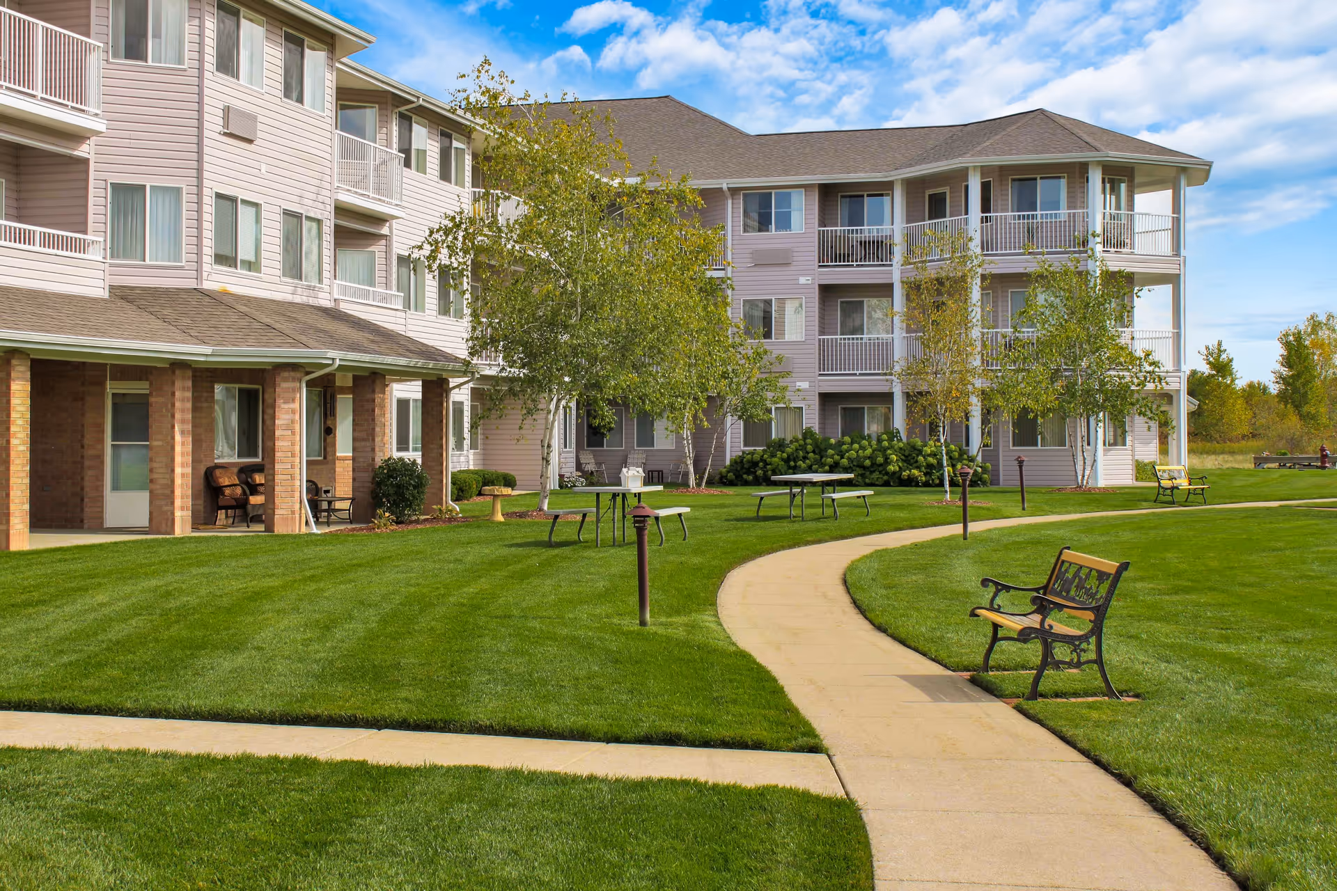 Outdoor view of Independence Village of Saginaw showing a well-maintained lawn with a curved concrete walkway, benches, picnic tables, and a three-story residential building with balconies and windows under a partly cloudy sky.