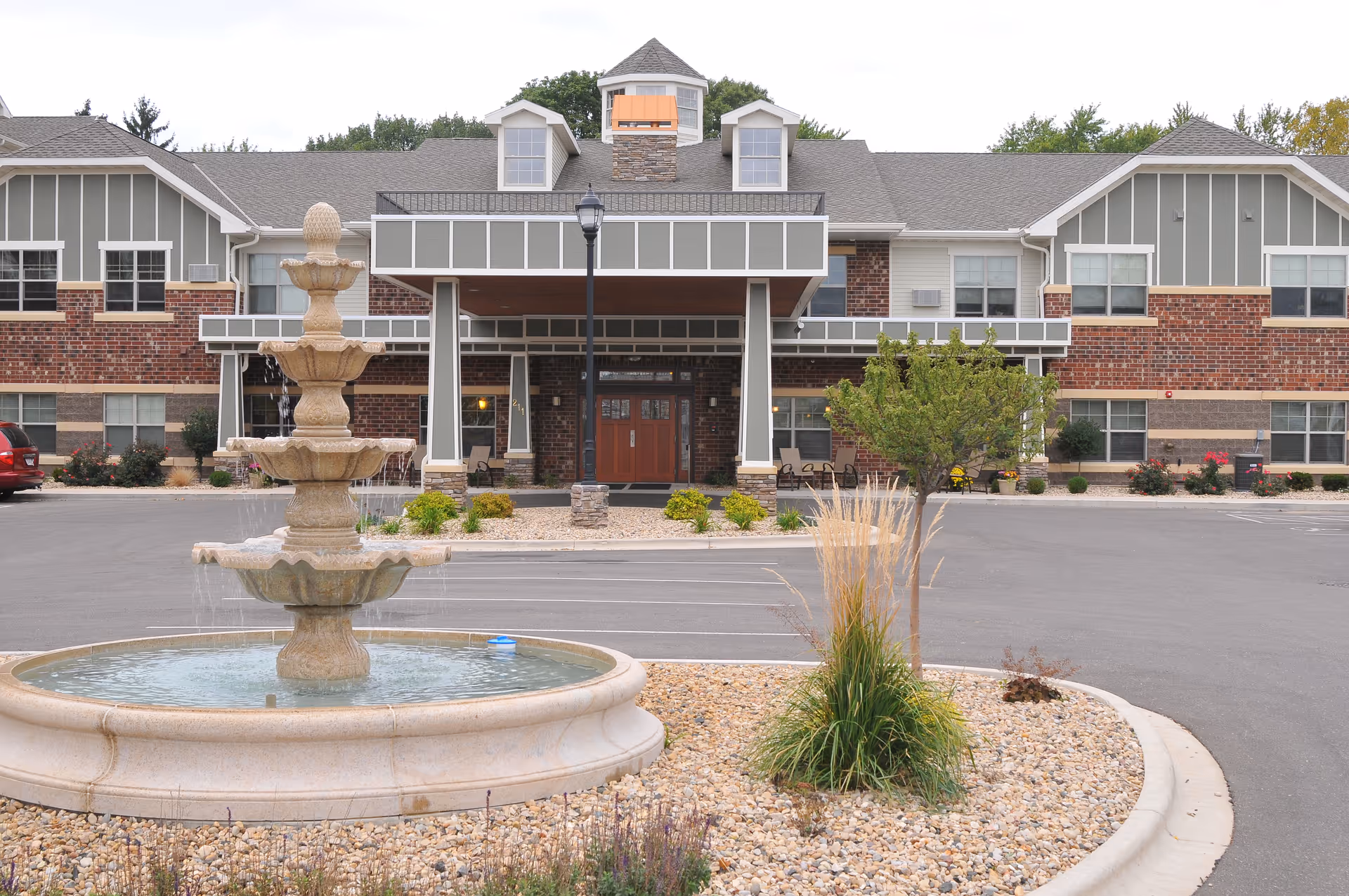 Tiered stone fountain and landscaped circular driveway in front of the entrance to a two-story brick-and-siding senior living building.