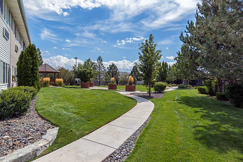A well-maintained outdoor garden area at Sierra Place Senior Living featuring a curved concrete walkway, green grass, various trees and shrubs, large planter pots, and a wooden gazebo under a partly cloudy blue sky.
