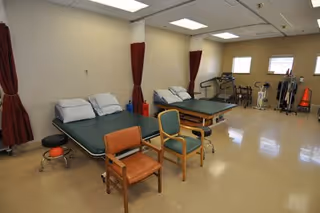 A rehabilitation room with two padded treatment tables, several chairs, exercise equipment including stationary bikes and weights, beige walls, and windows with red curtains.