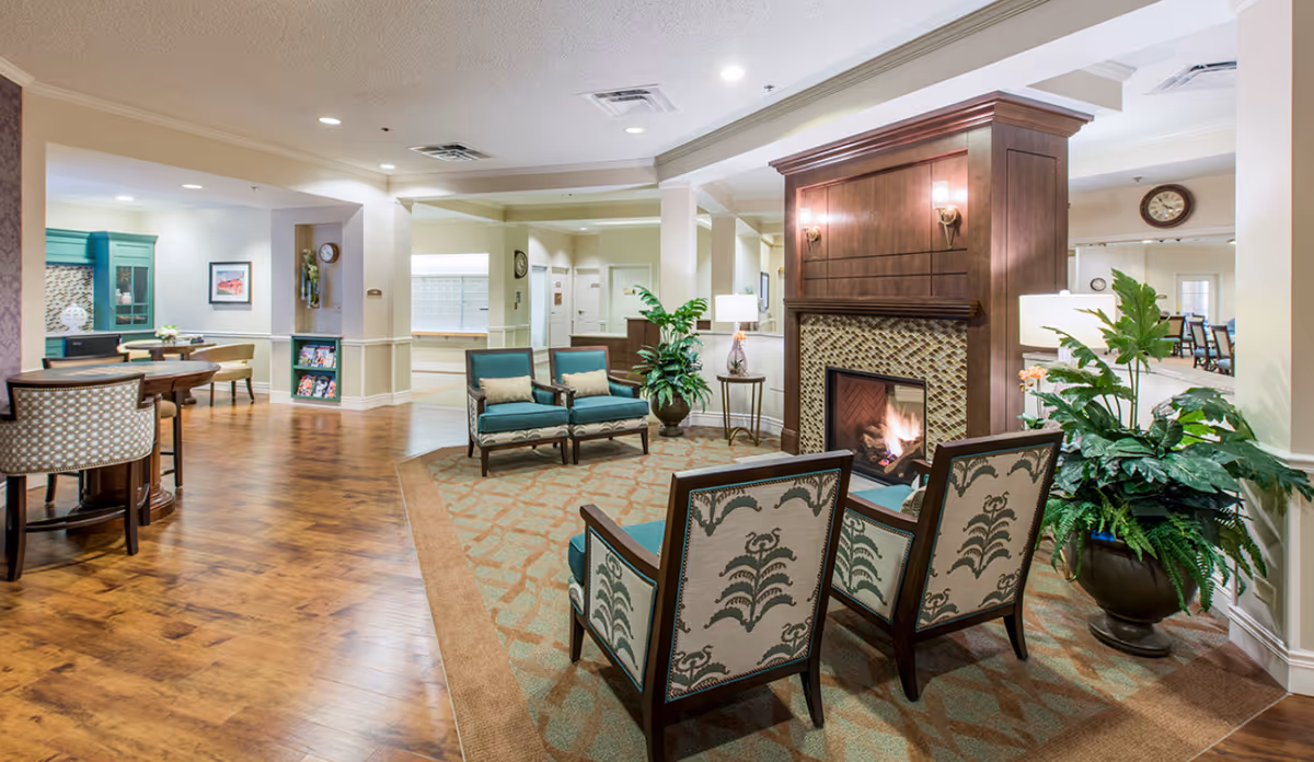 Comfortable communal lounge with a fireplace, chairs, tables and plants in a senior living facility.
