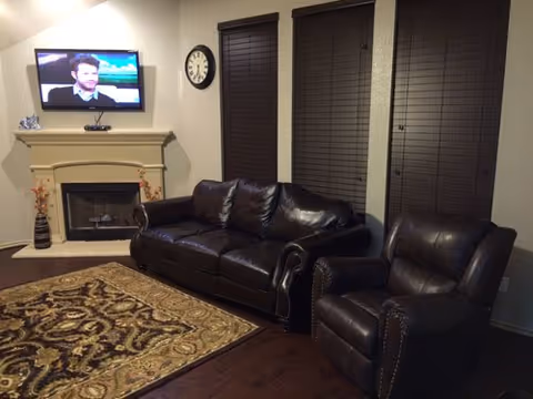 A cozy living room with a dark brown leather sofa and matching armchair, a patterned area rug on a wooden floor, a wall-mounted flat-screen TV above a fireplace, a clock on the wall, and two windows with closed dark blinds.