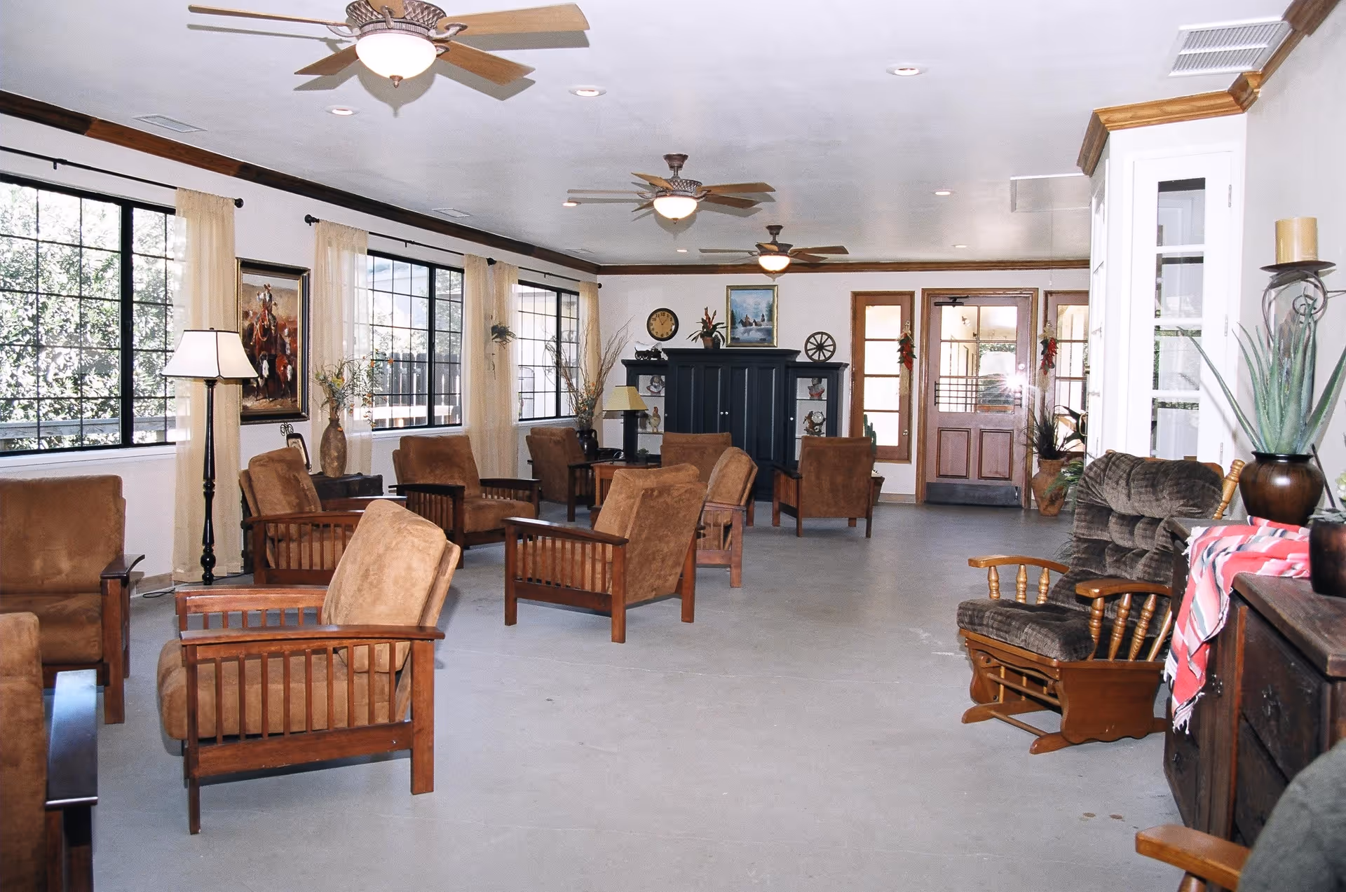Bright communal lounge with wooden armchairs, ceiling fans, large windows, and a doorway at the far end.