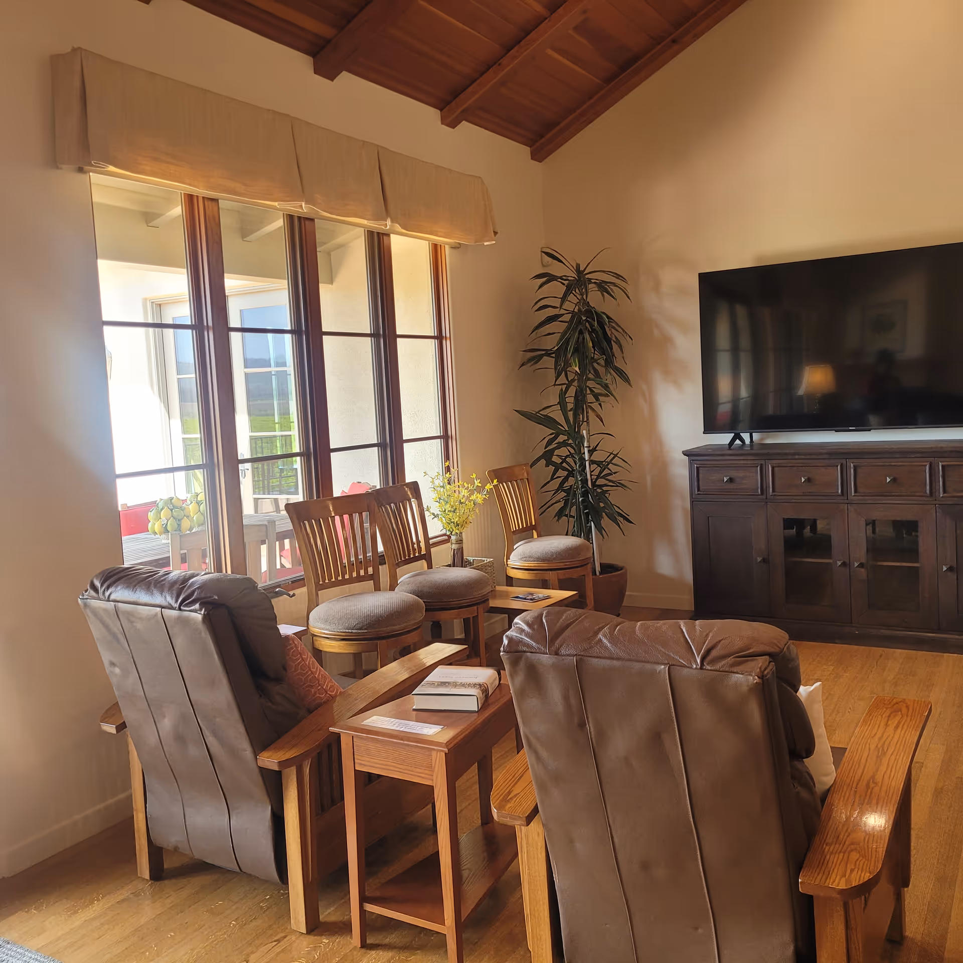 Sunlit living room with two leather recliners, wooden chairs and side tables facing a large TV on a dark wood console near tall windows.