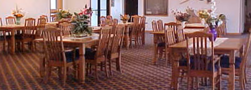 A spacious dining room with multiple wooden tables and chairs arranged neatly. Each table has a floral centerpiece, and the room is well-lit with natural light coming through the windows. The floor is carpeted with a patterned design.