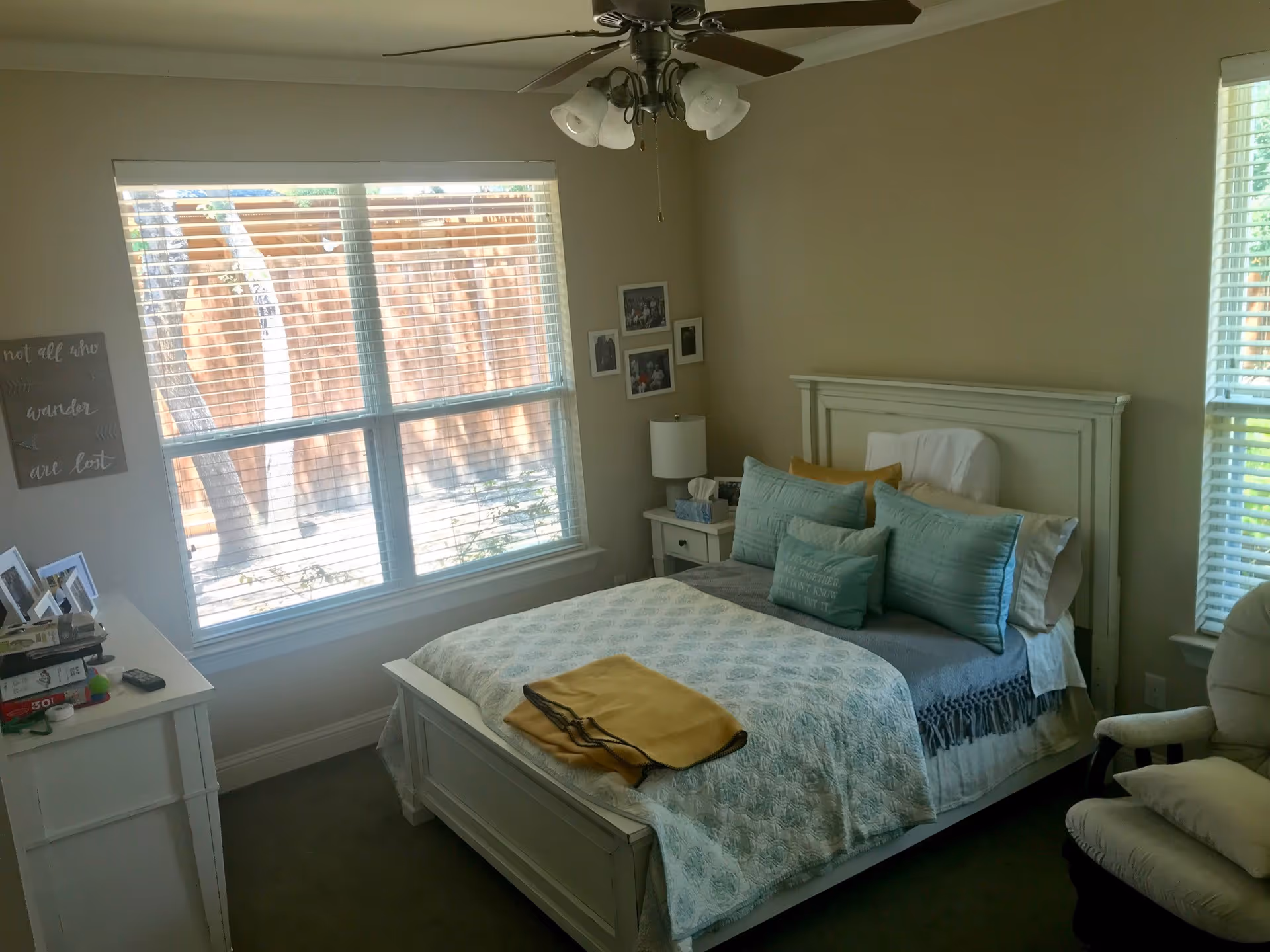 A cozy bedroom with a white wooden bed frame, a bed made with blue and white bedding and multiple pillows. There is a yellow blanket folded at the foot of the bed. The room has two windows with blinds allowing natural light to enter. A white nightstand with a lamp and tissue box is beside the bed. A white dresser with picture frames and other items is on the left side of the room. A ceiling fan with lights hangs from the ceiling. The walls are painted beige and decorated with framed photos and a wall art piece.