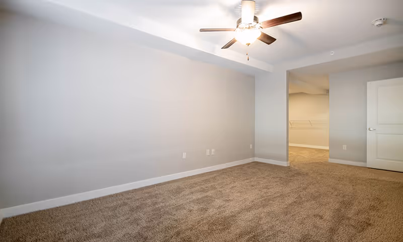 Empty room with beige carpet, light gray walls, a ceiling fan with light, and an open doorway leading to a walk-in closet with wire shelving.