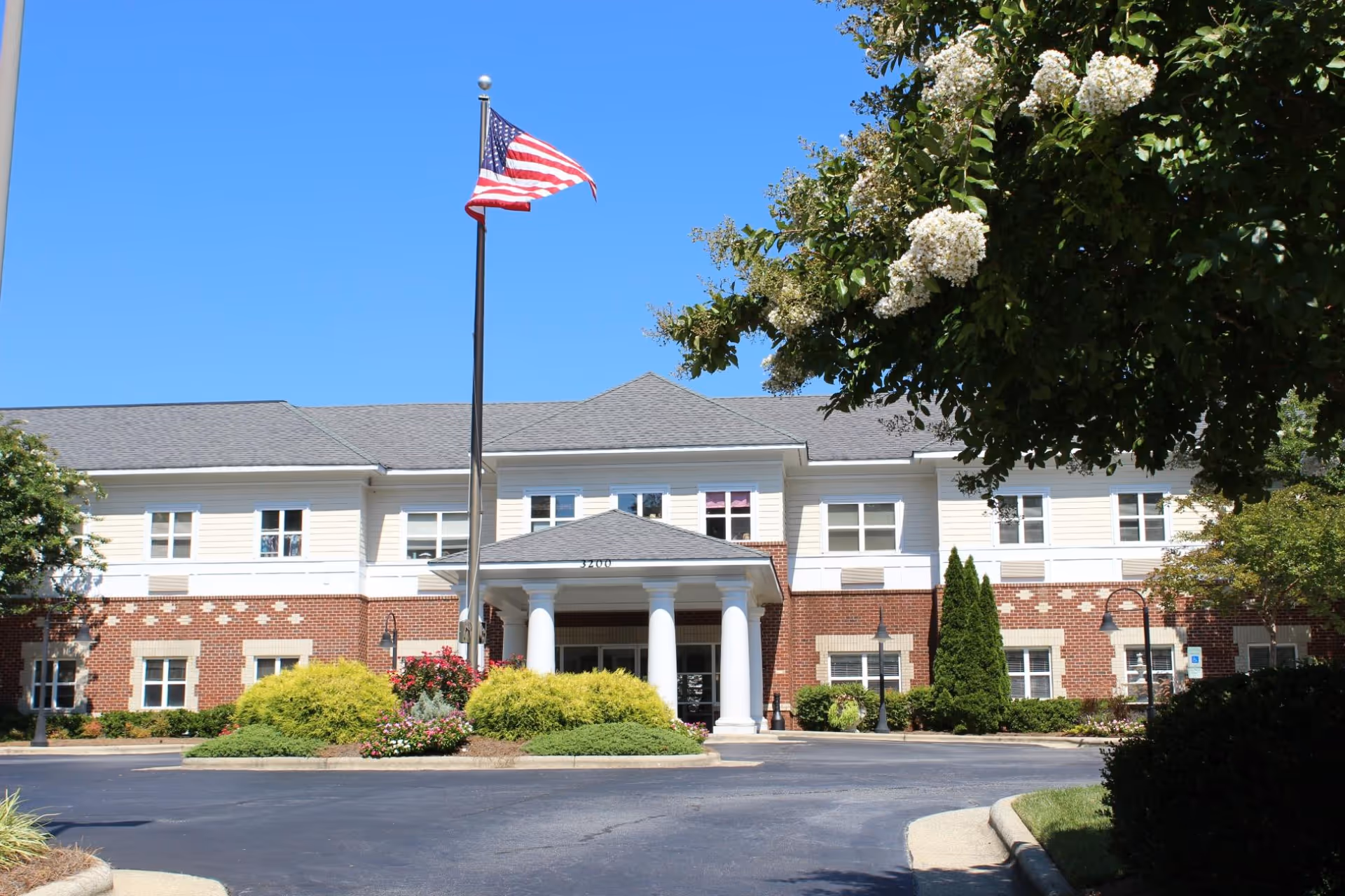 Front exterior of a two-story senior living building with a columned entrance, American flag on a pole, and landscaped driveway under a clear blue sky.
