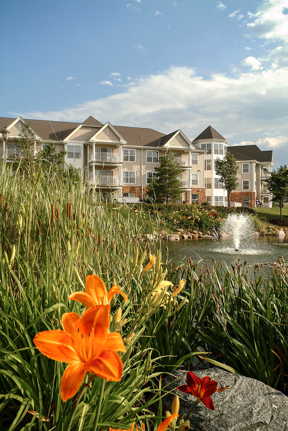 A multi-story cream-colored residential building by a pond with a fountain, surrounded by tall grasses and orange lilies under a blue sky.