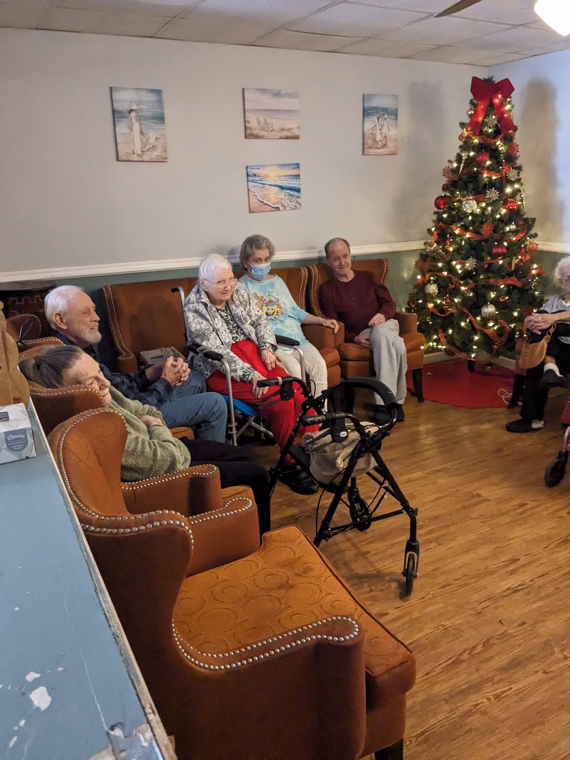 A group of elderly people sitting in a living room area with brown armchairs and a decorated Christmas tree with lights and ornaments in the corner. The room has wooden flooring and four beach-themed paintings on the wall. One person is wearing a face mask and another has a walker in front of them.