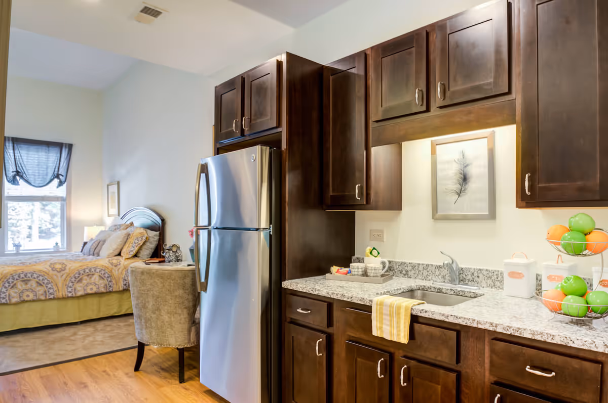 A cozy studio apartment featuring a kitchen area with dark wood cabinets, a granite countertop, a stainless steel refrigerator, and a sink with a yellow striped towel hanging on the edge. The kitchen counter has a tray with cups and snacks, two white canisters, and a two-tiered fruit basket with green and orange apples. In the background, there is a bed with patterned bedding, a window with a dark curtain, and a small upholstered chair next to a nightstand with a lamp and decorative items.