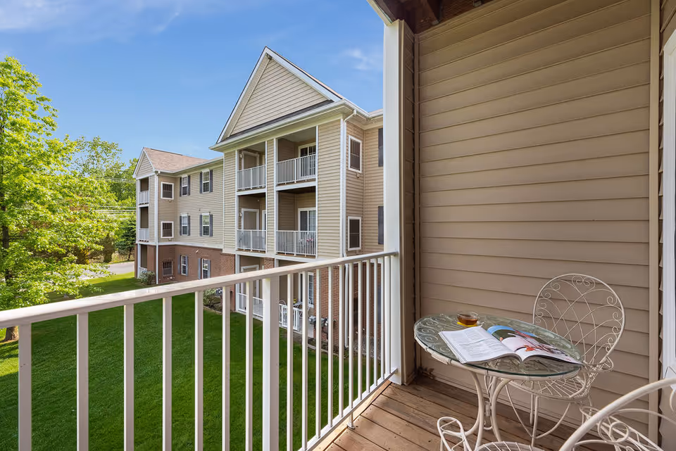 Balcony with a small glass-topped table and chair overlooking a grassy courtyard and neighboring apartment building.