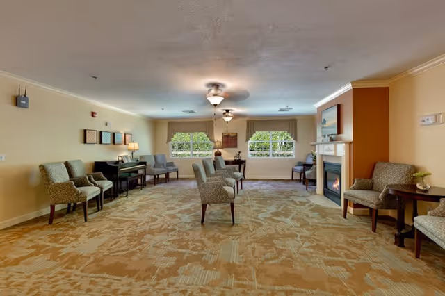 A spacious living room area with patterned carpet, multiple upholstered chairs arranged in pairs, a fireplace with a painting above it, two windows with beige curtains, a piano with a lamp on it, and ceiling fans with lights.