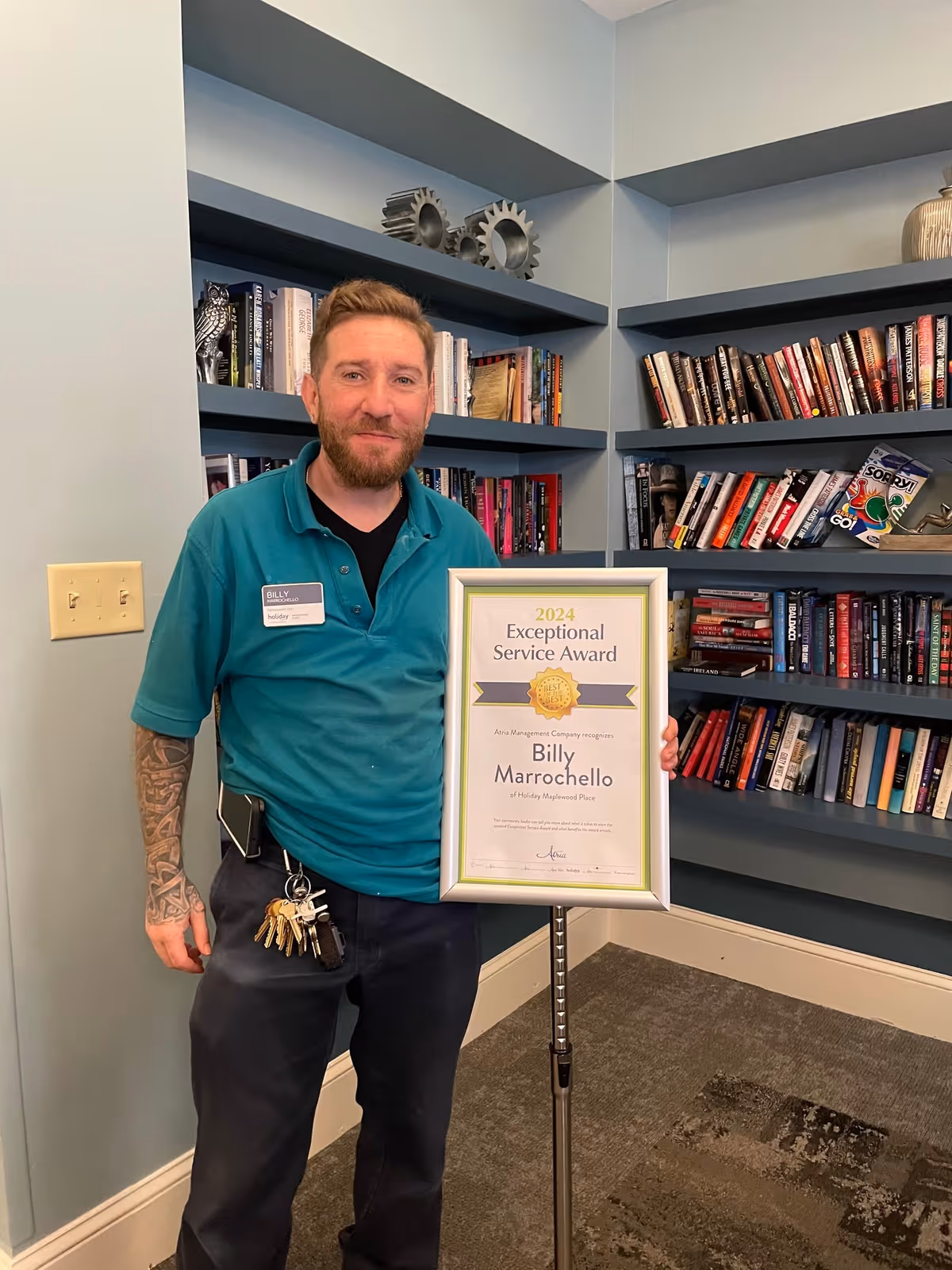 A man wearing a teal polo shirt and black pants stands indoors holding a framed certificate that reads '2024 Exceptional Service Award' presented to Billy Marrochello at Holiday Maplewood Place. Behind him are blue built-in bookshelves filled with books and decorative items.