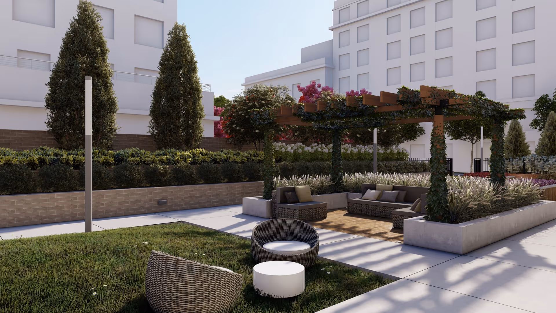Outdoor seating area in a senior living facility with wicker chairs and a small round table on grass, and a shaded pergola with cushioned sectional seating surrounded by plants and greenery, with a multi-story building in the background.