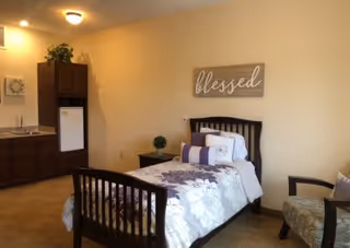 A cozy bedroom with a single bed featuring floral bedding and multiple pillows. Above the bed is a wall decoration with the word 'blessed'. To the left, there is a small kitchenette area with a sink, cabinets, and a mini refrigerator. A cushioned armchair is positioned to the right of the bed. The room has warm lighting and a neutral color scheme.