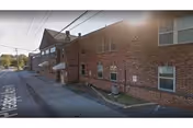 Exterior view of a brick building along a street with power lines overhead. The building has multiple windows and a small awning over a side entrance. The street is empty and there is some greenery visible in the distance.