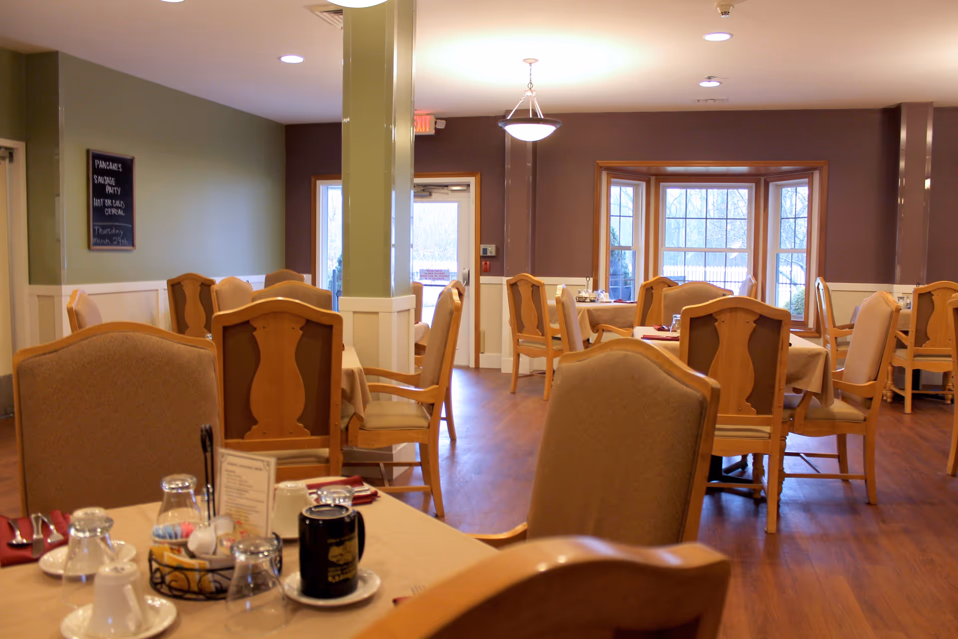 Dining room with multiple set tables and wooden chairs, large windows, and warm lighting.