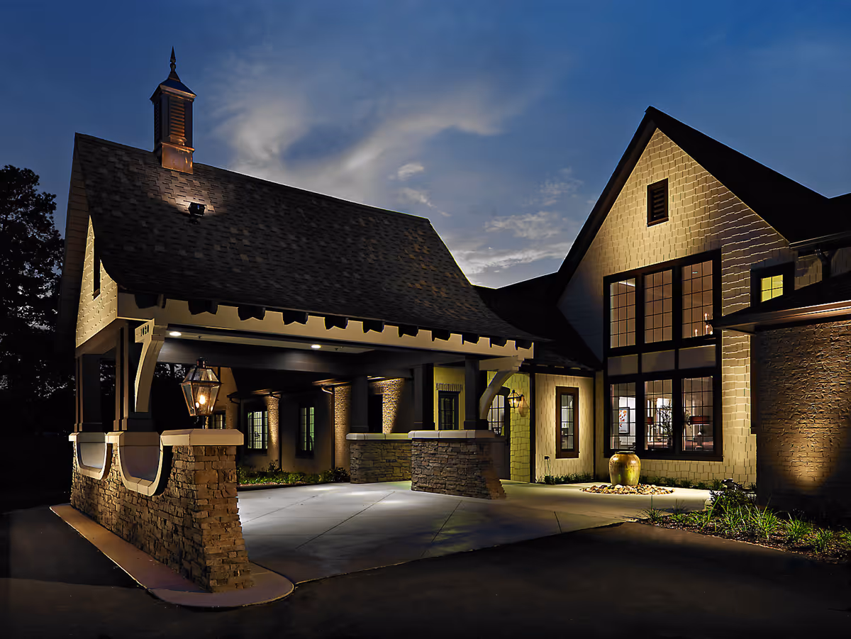 Exterior view of Westminster Assisted Living facility at dusk, showing a covered entrance with stone pillars and warm lighting illuminating the building's facade and large windows.