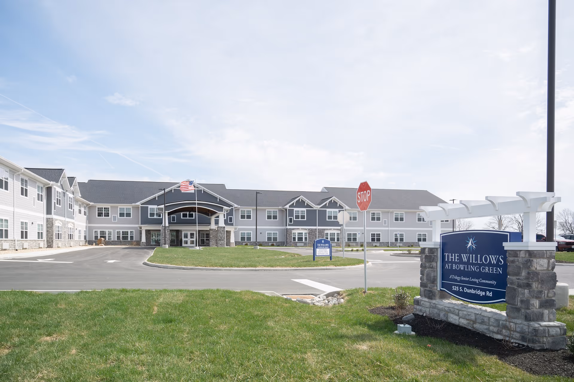 Exterior view of The Willows at Bowling Green senior living community building with a driveway, an American flag, a stop sign, and a stone sign displaying the facility name and address.