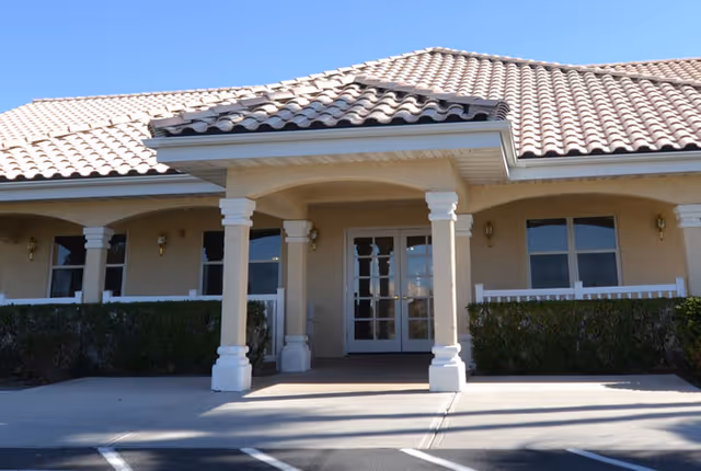 Exterior front entrance of a single-story building with beige stucco walls, a tiled roof, white columns supporting a covered porch, and glass double doors. There are bushes along the front and a parking area in front of the entrance.