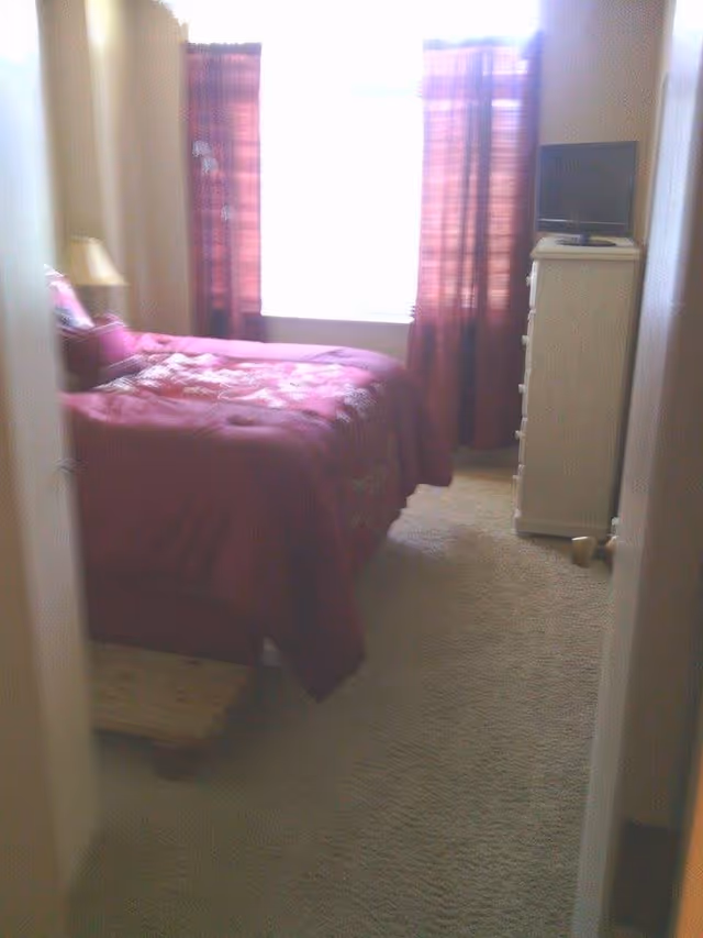 A small bedroom with a bed covered in a red and floral patterned comforter, a wooden step stool at the foot of the bed, a nightstand with a lamp on the left side, and a white dresser with a small TV on top near a window with red curtains.