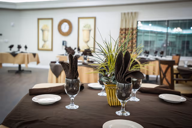 A dining room with tables covered in brown tablecloths, each set with white plates, glasses with folded brown napkins, and a small plant centerpiece. The room has framed artwork on the walls and large windows with blinds.