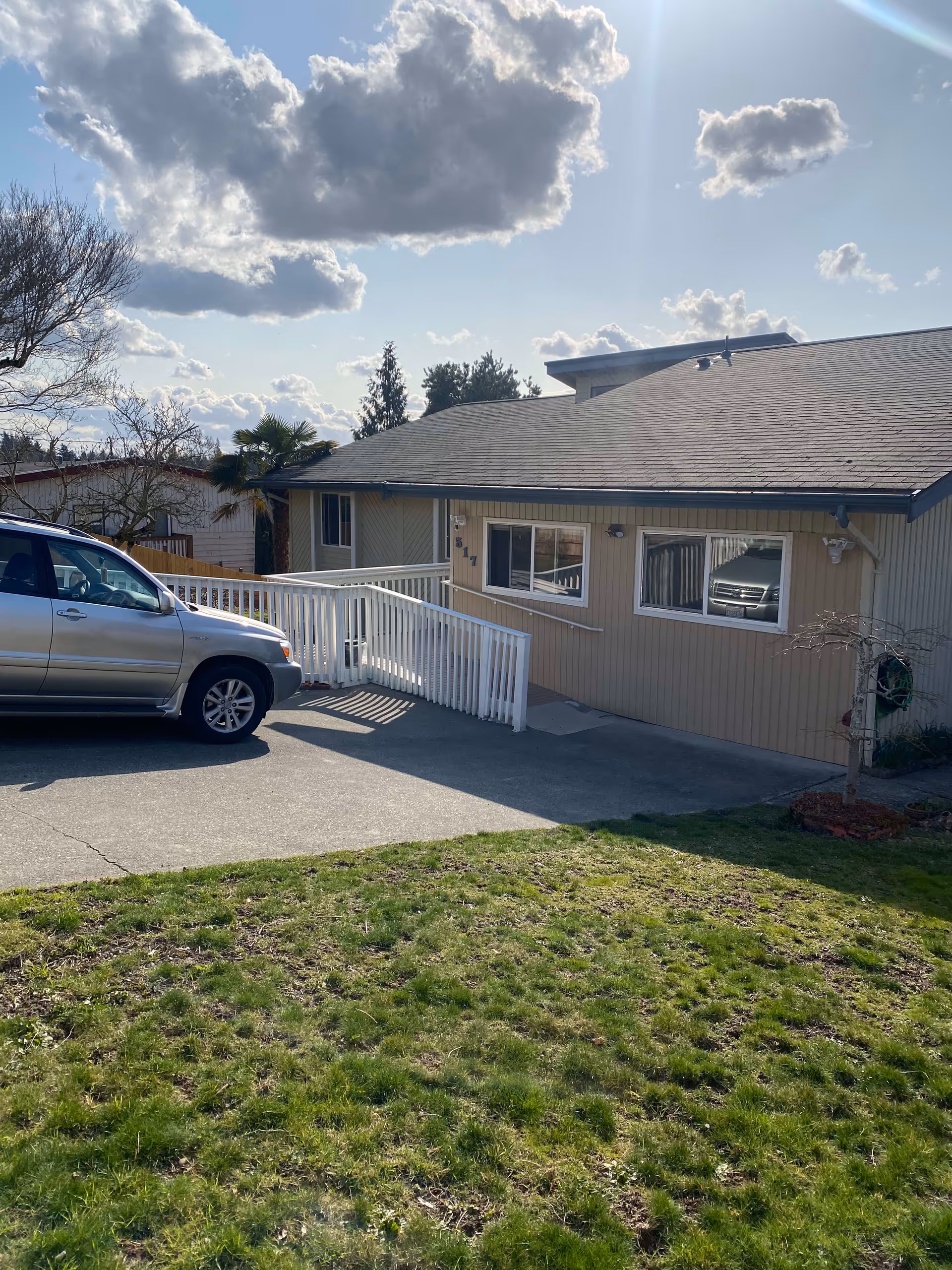 Single-story beige house with a front wheelchair ramp, a parked SUV, and a grassy lawn under a cloudy sky.