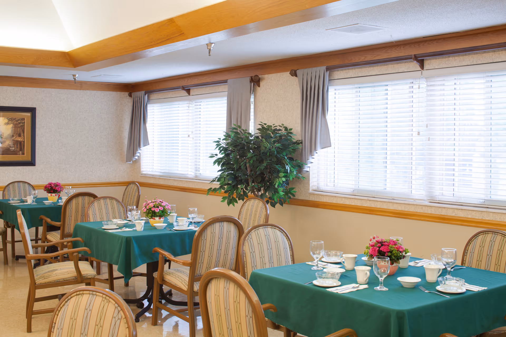 A dining room with several tables covered in green tablecloths, each set with plates, cups, glasses, and silverware. There are cushioned wooden chairs around the tables, a potted plant near the windows, and framed artwork on the wall. The room has large windows with blinds and curtains, and a ceiling with wooden beams.