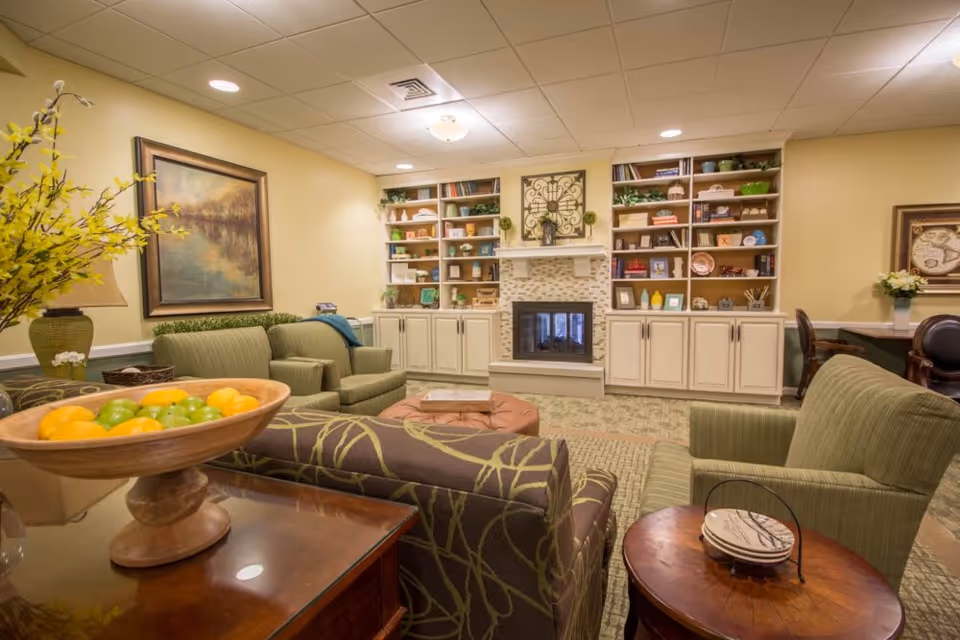 A cozy living room area with green upholstered chairs and a patterned sofa arranged around a central ottoman. There is a wooden bowl with green and yellow apples on a table in the foreground. The back wall features built-in shelves filled with decorative items and books, and a fireplace with a tiled surround. The room is warmly lit with recessed ceiling lights and has beige walls and carpeted flooring.