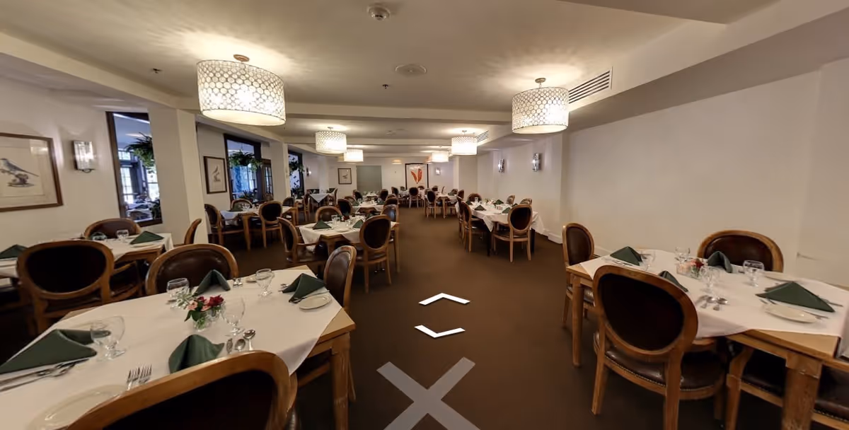 Large formal dining room with multiple tables set with white tablecloths, green napkins, and wooden chairs under pendant lights.