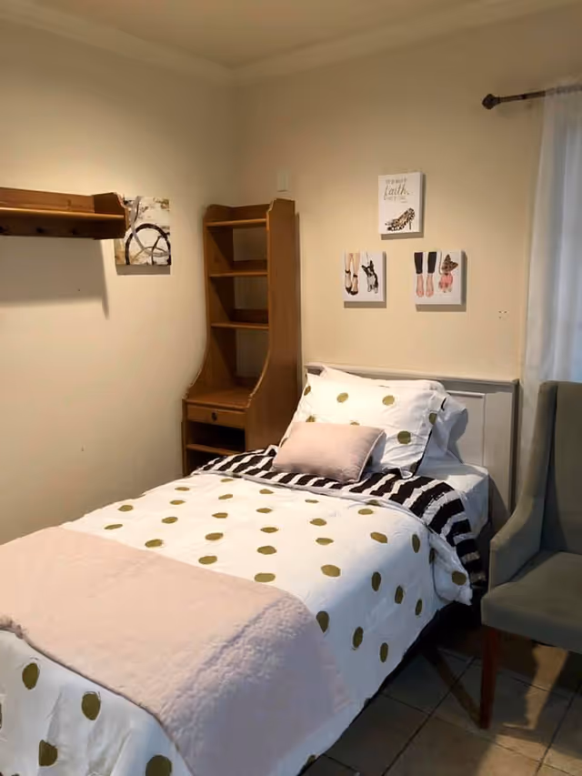 A small bedroom with a single bed covered in white bedding with gold polka dots and a light pink blanket at the foot. There is a light pink pillow and a black and white striped blanket on the bed. Next to the bed is a wooden shelving unit with a small desk area. On the wall above the bed are three small framed pictures, and to the right is a window with a sheer white curtain. A gray upholstered chair is positioned near the bed.