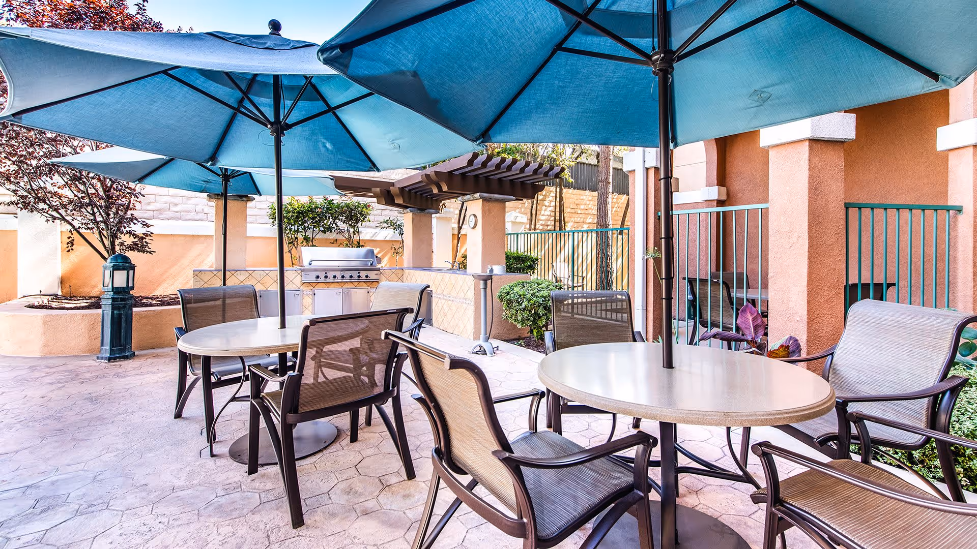 Outdoor patio area with round tables and chairs under large blue umbrellas. There is a built-in grill and a pergola in the background, surrounded by plants and a stone wall.