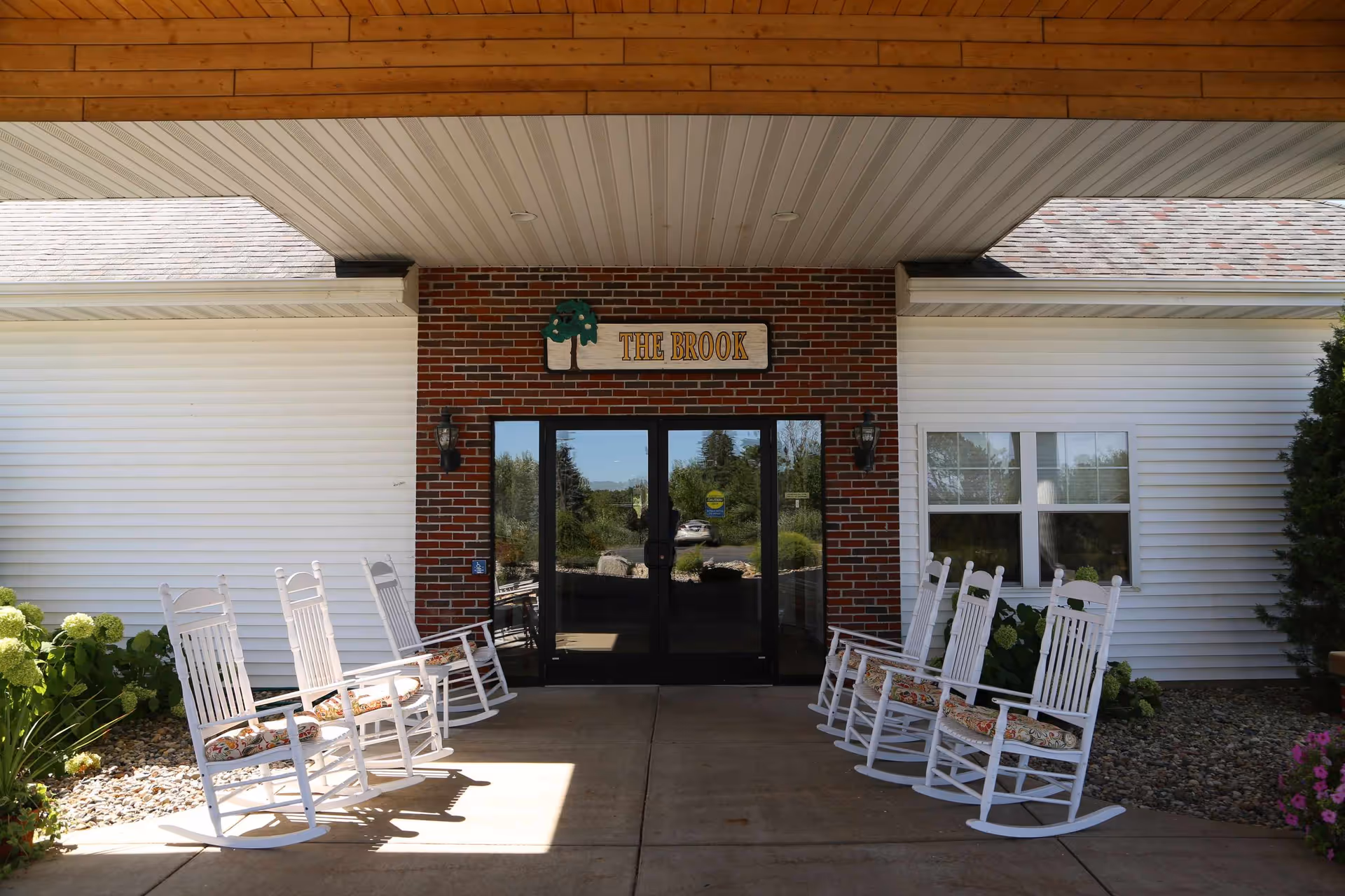 Entrance of The Brook senior living facility with a covered porch area featuring six white rocking chairs with floral cushions, two on the left and four on the right. The building has white siding with a brick section around the glass double doors. A sign above the doors reads 'THE BROOK' with a tree graphic. There are plants and flowers on either side of the entrance.