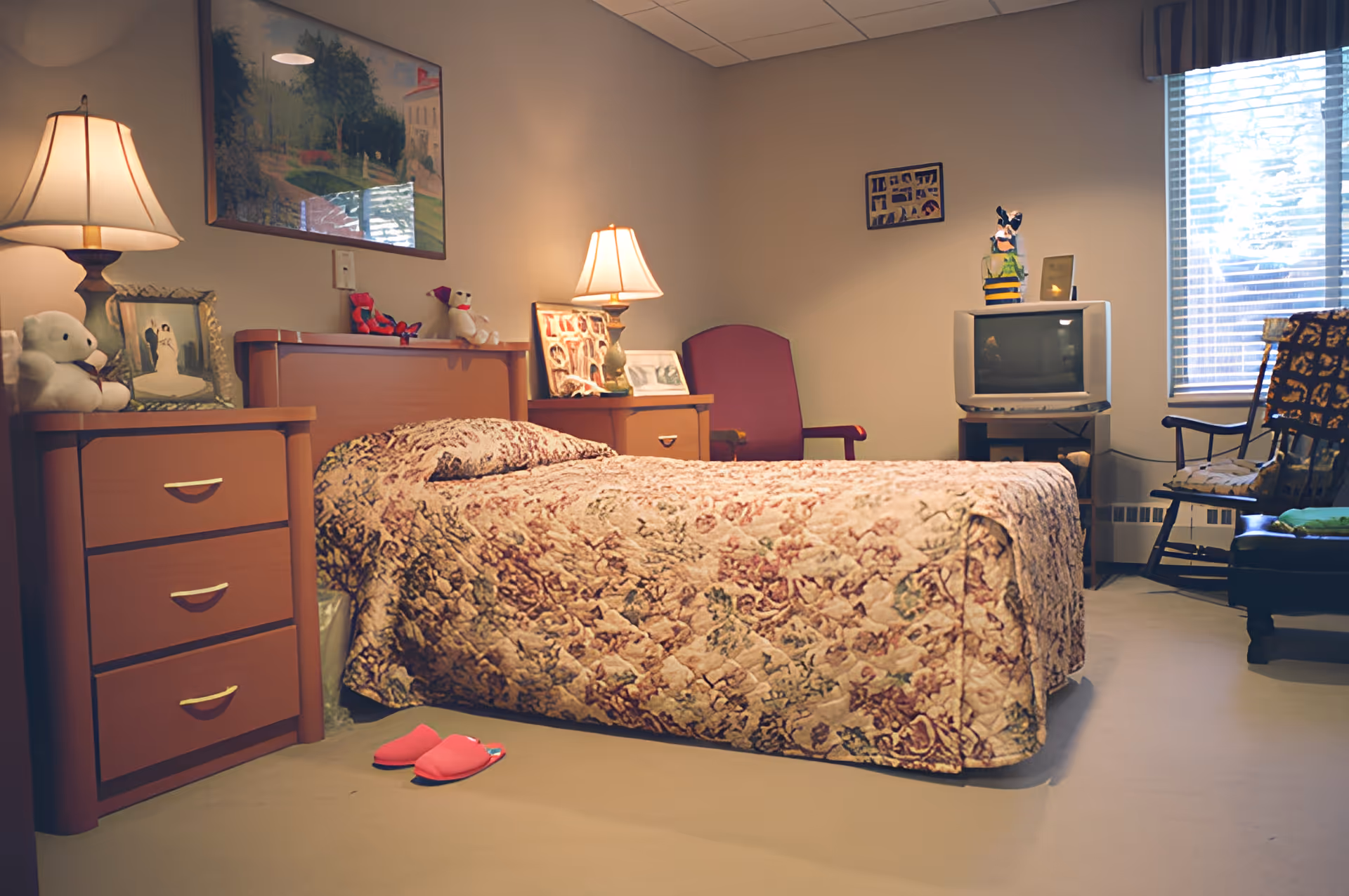 A cozy bedroom with a floral bedspread, nightstands and lamps, a TV, chairs by a window and pink slippers on the floor.