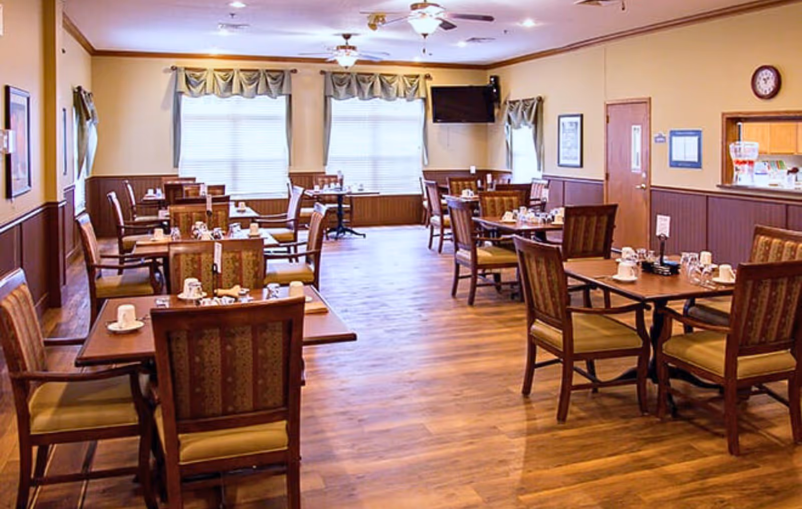 A dining room with several wooden tables and chairs arranged neatly. Each table is set with cups, glasses, and napkins. The room has wooden flooring, beige walls with dark wood paneling, and windows with curtains allowing natural light to enter. A wall-mounted TV and a clock are visible on the walls.